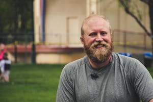 A cheerful bearded man smiling outdoors in a casual setting with greenery.
